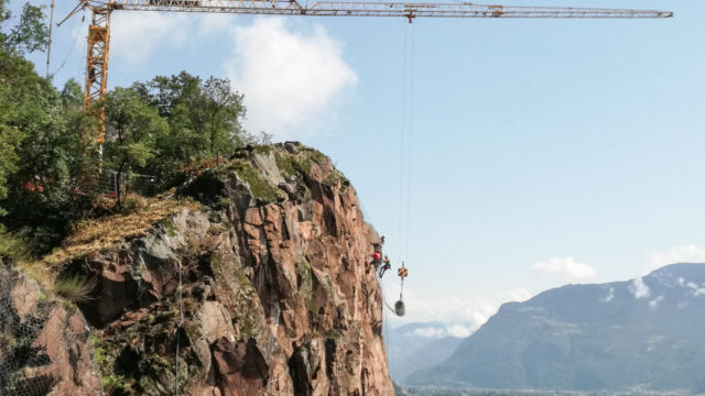 Messa in sicurezza di pareti rocciose tramite rete zincata e pannelli in funi di acciaio: Installazione della rete zincata sulla parete rocciosa con l'uso della gru. Laimburg, Vadena (BZ). Messa in sicurezza di pareti rocciose tramite rete zincata e pannelli in funi di acciaio: Installazione della rete zincata sulla parete rocciosa con l'uso della gru. Laimburg, Vadena (BZ).